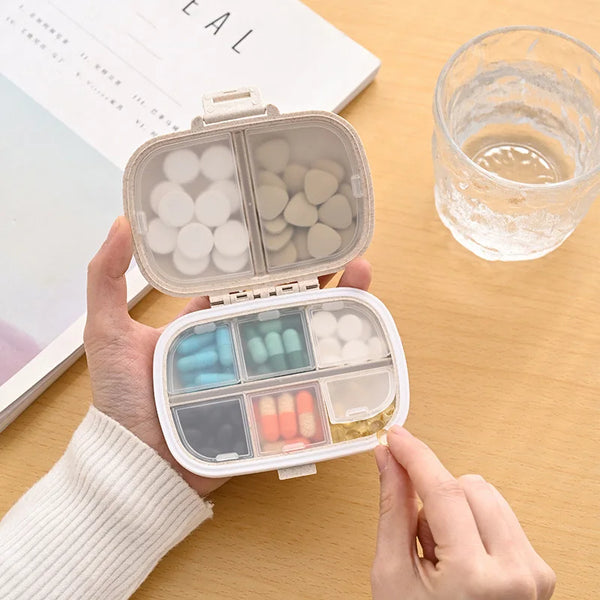 A hand holds an open weekly pill organizer with eight compartments containing various colored pills on a wooden surface.