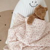 A soft floral muslin baby blanket with a white quilted background and a brown teddy bear on a light beige surface.