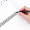 A hand holds a black pencil against a stainless steel precision straight ruler on a white sheet of paper.