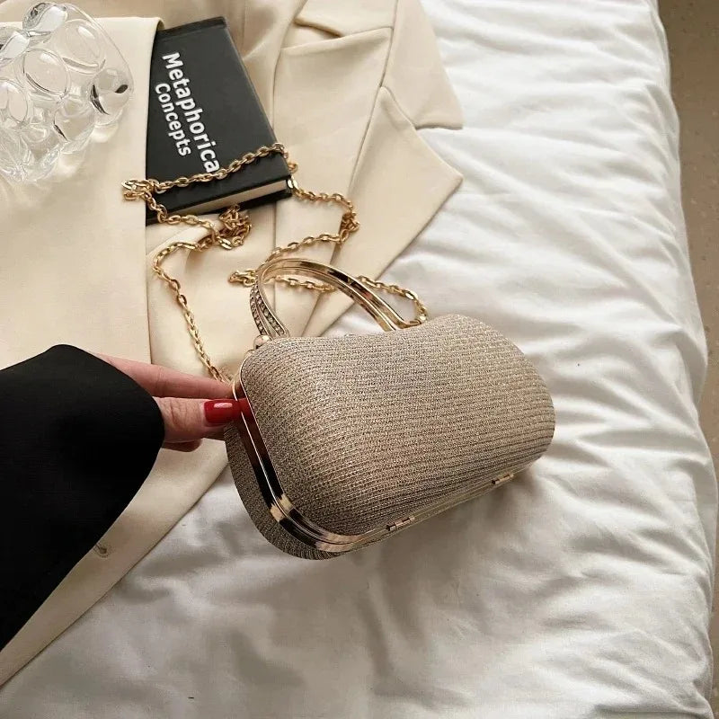A beige envelope clutch with a gold chain strap resting on white bedding next to a book.