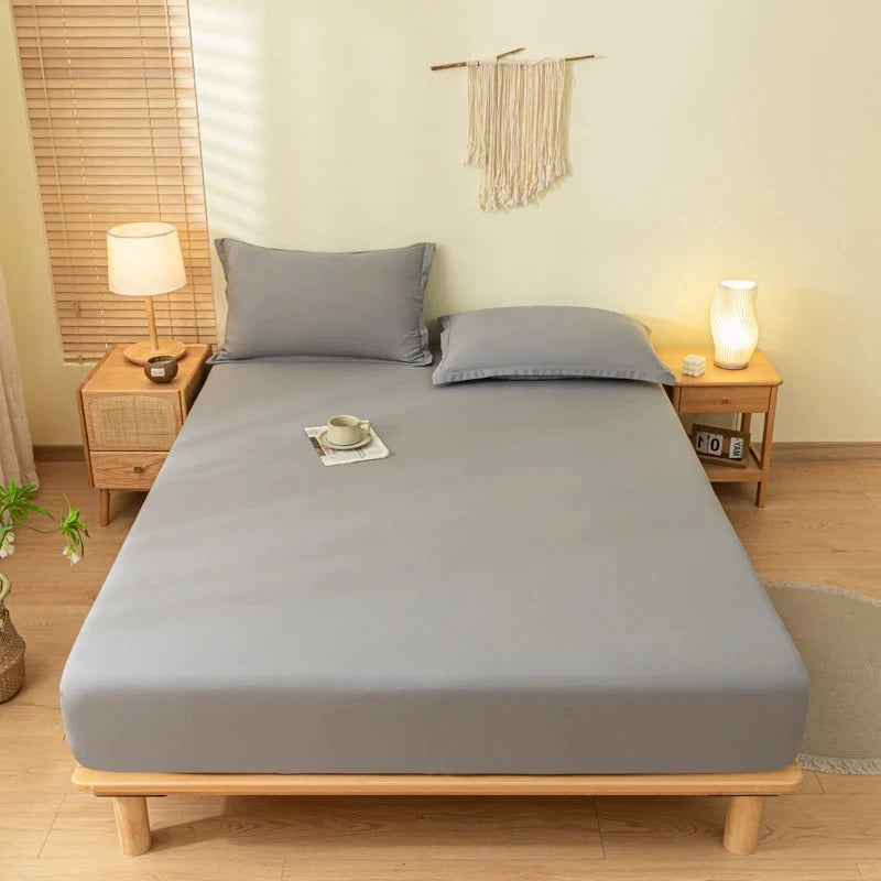 A light gray fitted bed sheet set with pillowcases is displayed on a wooden bed frame in a bright room.