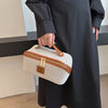 Eine Hand hält eine beige Reise-Make-Organizer-Tasche mit einem braunen Lederriemen vor einem schwarzen Kleid.