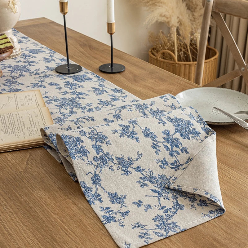 A blue and white floral gingham table runner laid on a wooden table with candles and dried grasses in the background.