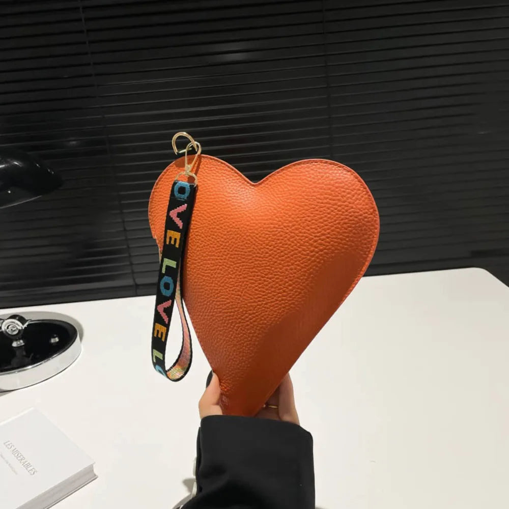 An orange heart-shaped PU coin purse with a black wrist strap is held up against a dark background.