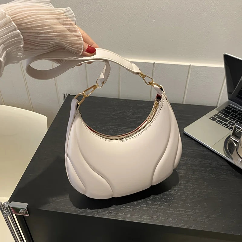 A white textured crossbody bag with gold hardware is held by a hand on a dark surface.