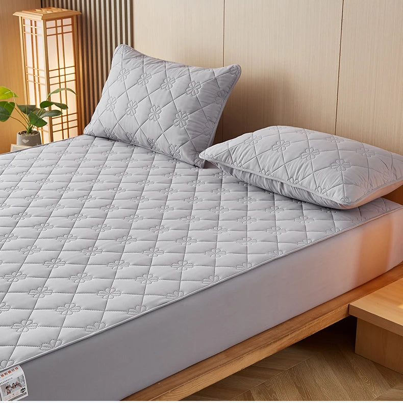 A gray quilted mattress protector is shown on a bed, with two pillows resting on top.