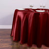 A round table set with a deep red satin tablecloth, white plates, and silverware, set on a wooden floor.