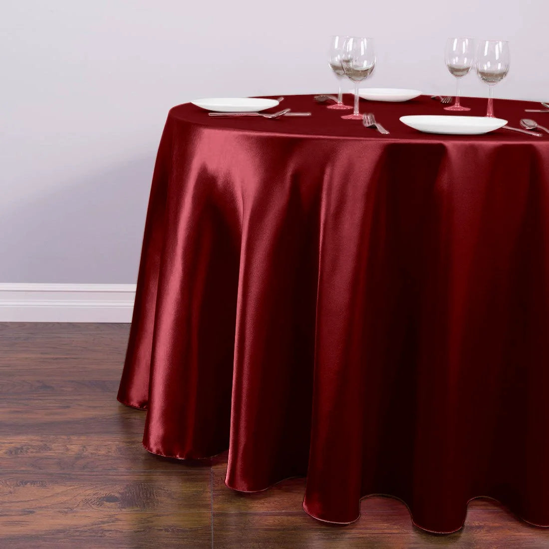 A round table set with a deep red satin tablecloth, white plates, and silverware, set on a wooden floor.
