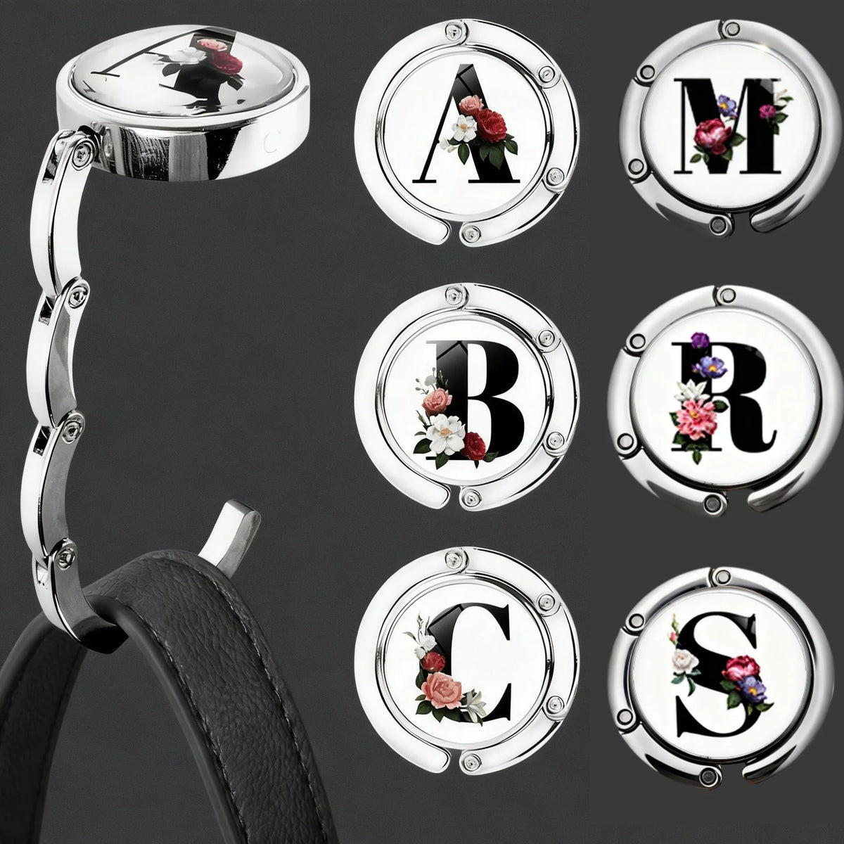 Silberne personalisierte Briefhaken mit den Buchstaben A, M, B, R, C und S auf einem schwarzen Lederriemen.