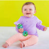 A smiling baby wearing a purple long-sleeved top and pink pants holding a green rattle toy.