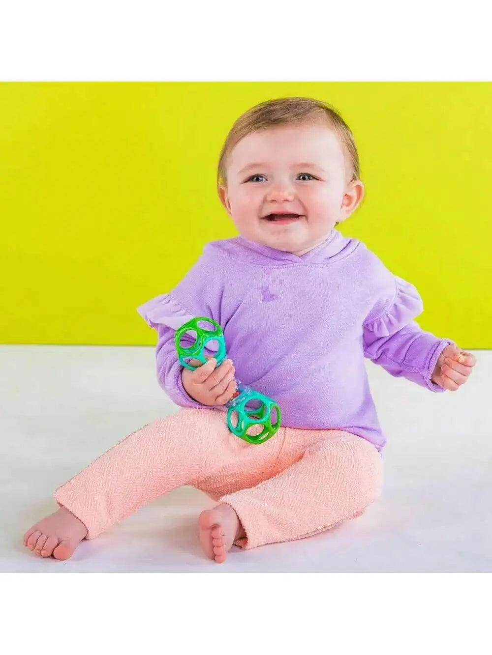 A smiling baby wearing a purple long-sleeved top and pink pants holding a green rattle toy.