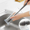 A hand is using a silicone bottle brush with a long handle to clean items in a stainless steel sink.