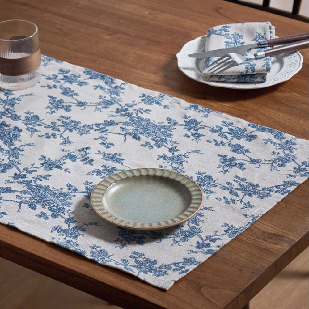 A light blue and white floral patterned table runner on a wooden table with silverware and a plate set nearby.