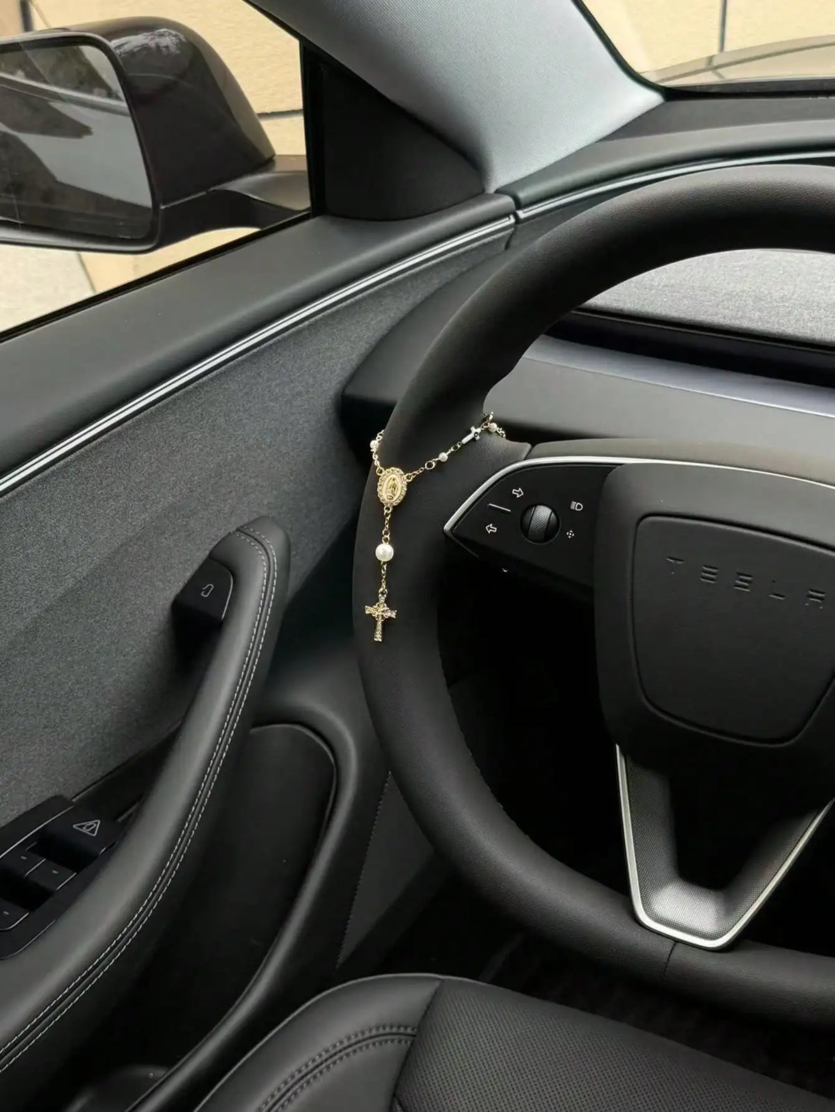 Interior view of a Tesla steering wheel showing a gold rosary pendant hanging from the wheel.
