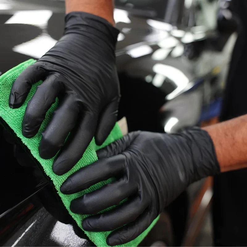A hand wearing a black nitrile glove is cleaning a dark surface with a bright green cleaning cloth.