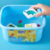 A hand places a green plastic dinosaur figure into a blue plastic container of Water Spirit craft materials.