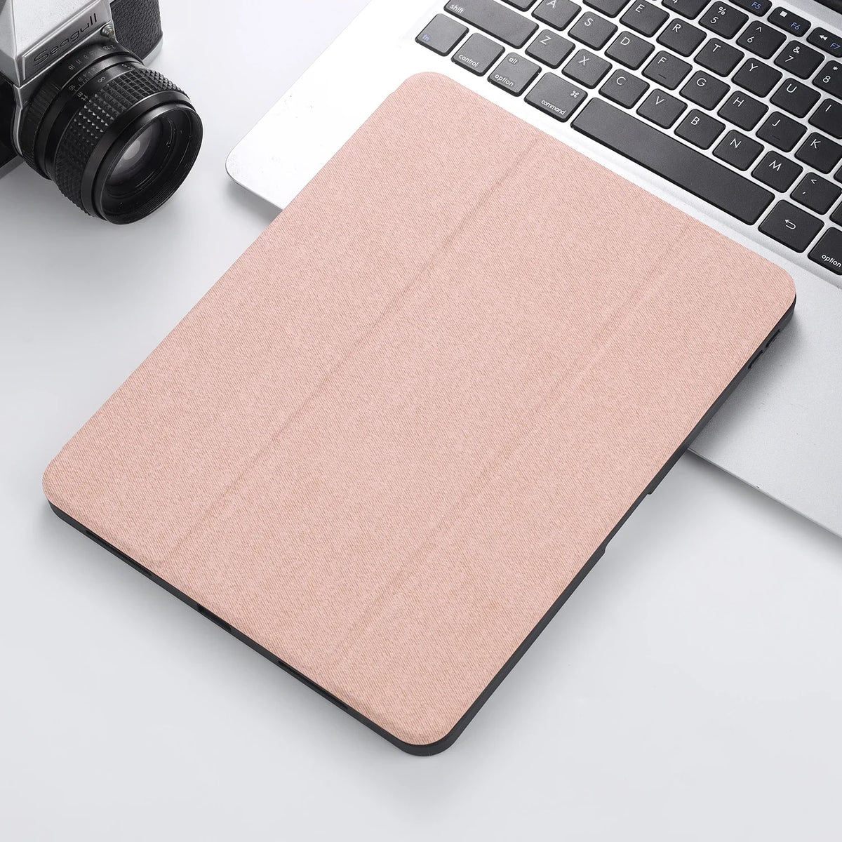 A light pink retro marble case for an iPad Pro 12.9 with a built-in pencil holder, placed next to a laptop and camera.