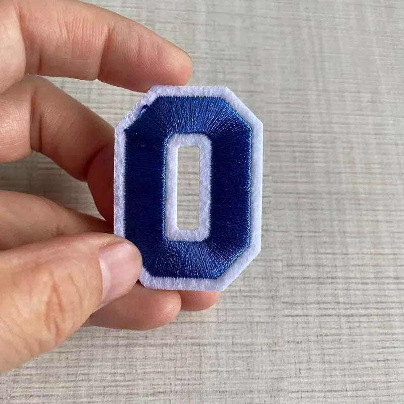A hand holds a dark blue felt iron-on 3D number patch shaped like the digit zero.