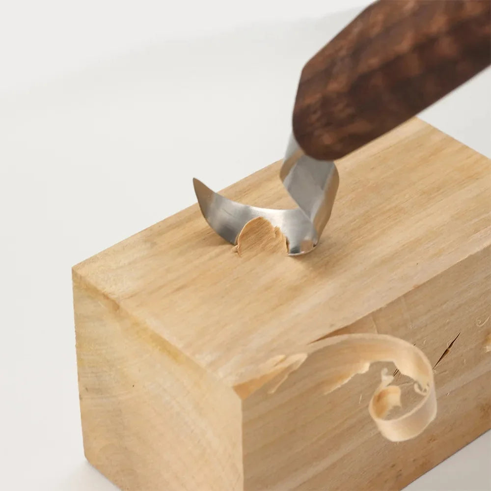 A hand using a metal carving tool with a wooden handle to carve a piece of wood.