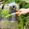A hand holds a carbon steel manual weeding tool set with a sickle, weeder, and hand shovel near a small water feature.