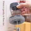 A hand is pressing a black dish brush with a built-in soap dispenser to allow liquid cleaning to flow out.