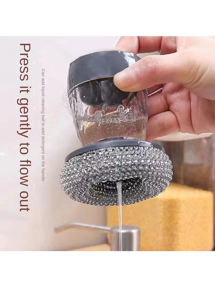 A hand is pressing a black dish brush with a built-in soap dispenser to allow liquid cleaning to flow out.