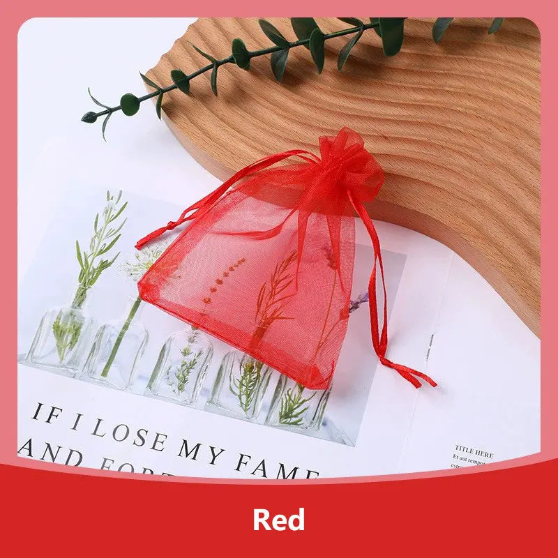A red organza drawstring gift bag tied with a ribbon, resting on a wooden surface next to small green plant sprigs.