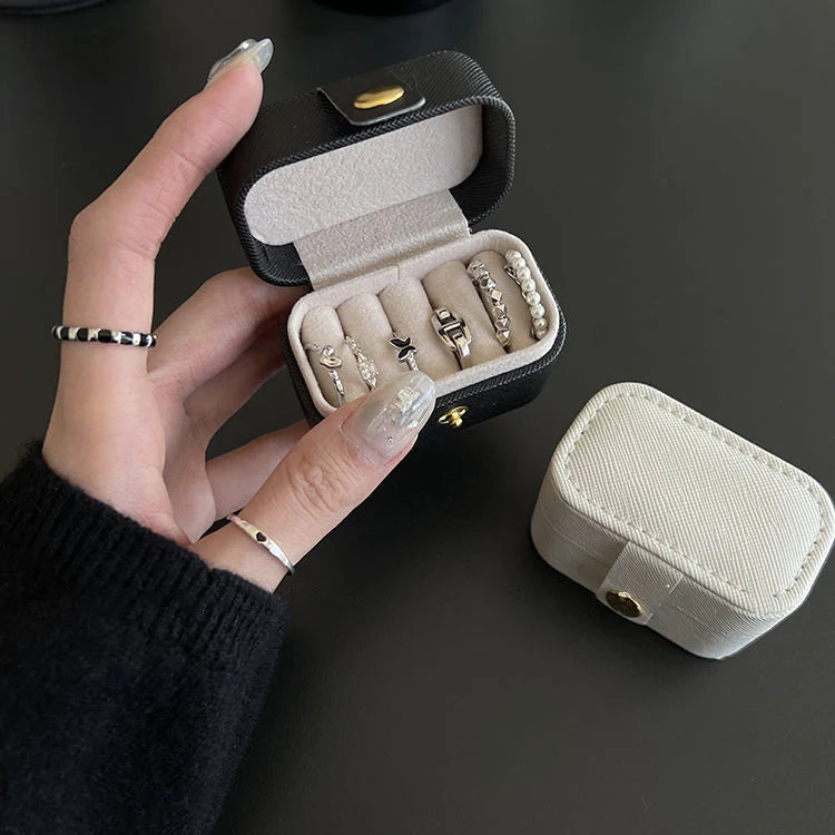A hand opens a compact black leather jewelry travel case revealing several silver rings and a small stud earring.