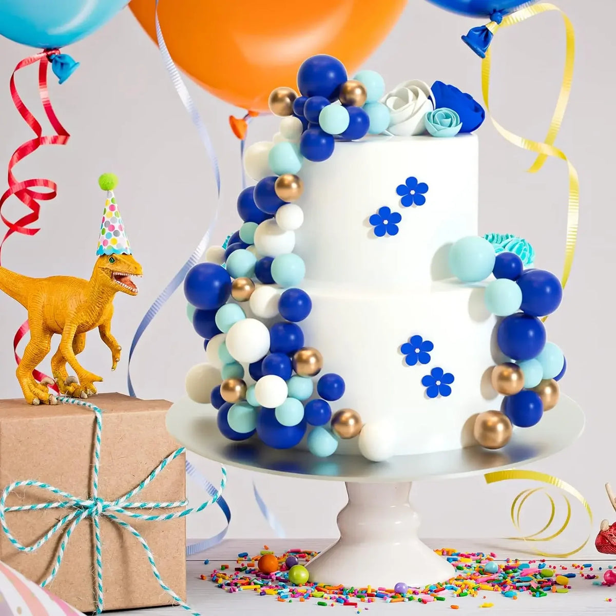 Ein weißes mehrstöckiges Kuchenmodell, dekoriert mit blauen und goldenen Kuglebonbons und Blumen, neben einem gelben Dinosaur