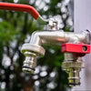 A close-up of a brass garden water faucet with a red lever handle and silver finish, set against a blurred outdoor background