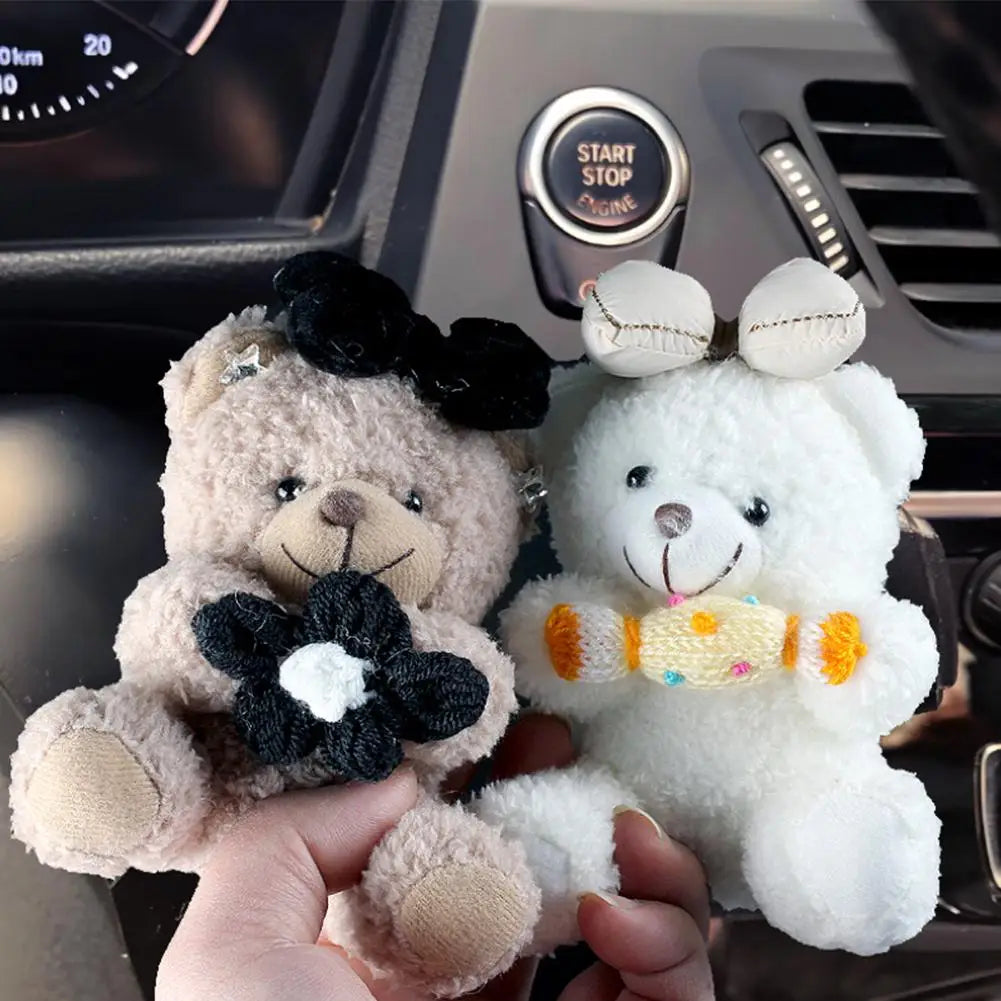 A hand holds a white plush bear toy next to a brown plush bear toy on a car gear shift cover.