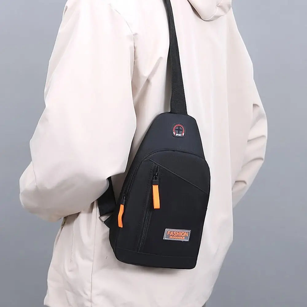 A black Oxford canvas crossbody chest bag with an orange strap and a small patch on the front, worn by a person in a light-co