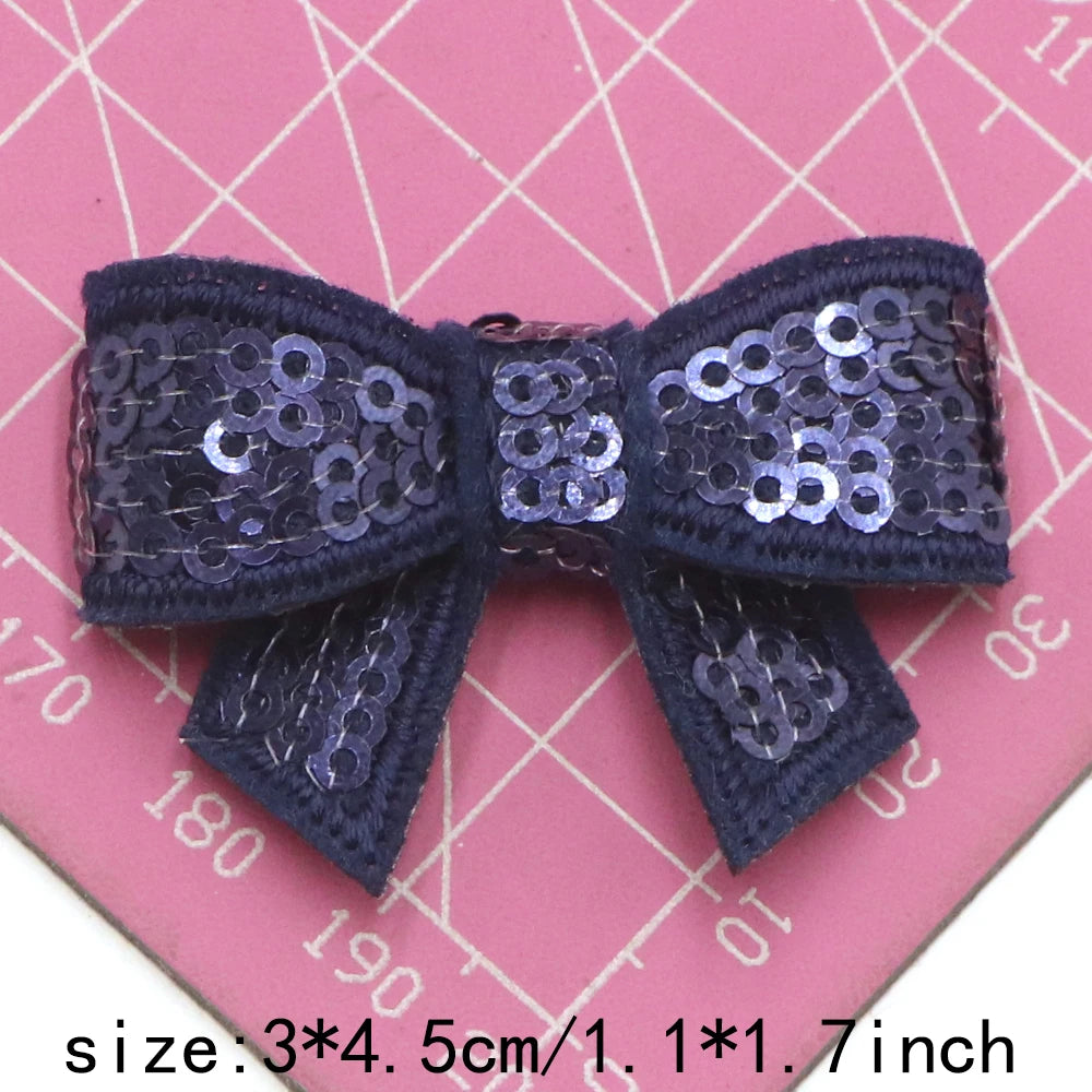 A dark purple sequin bow patch for DIY crafts shown against a pink grid background.