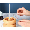 A hand is using a stainless steel beading needle to thread beads onto a wooden tool.