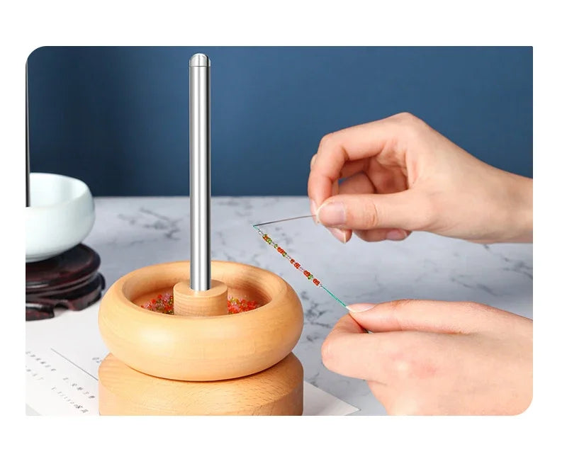 A hand is using a stainless steel beading needle to thread beads onto a wooden tool.