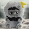 A soft gray plush toy resembling a face with black features and yellow ear, sitting on a light fabric.