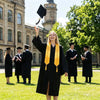 A smiling woman in a black graduation gown and yellow hood raises her hand in celebration outdoors.