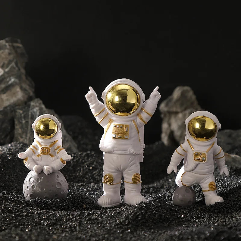 Three handcrafted astronaut figurines in white and gold resin standing on dark gravel.