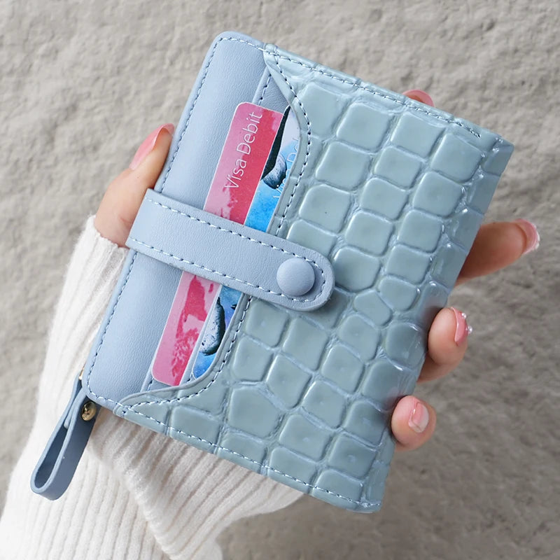 A hand holds a light blue crocodile-embossed clutch wallet with a grey leather strap.