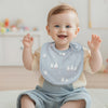 A smiling baby wearing a light blue cotton bib with white flocking, sitting on a white rug.