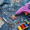 A purple hot melt glue gun is shown with various craft supplies like beads, gems, and glue sticks on a denim background.