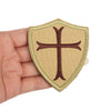 A beige shield-shaped patch embroidered with a dark brown cross, held in a hand.