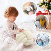 A baby is holding a soft, cream-colored knitted or velvet throw pillow on a white surface.