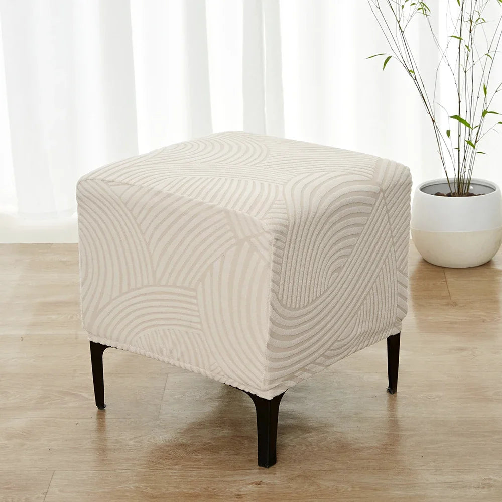 A beige geometric jacquard stretch ottoman cover placed on a wooden floor near a window.