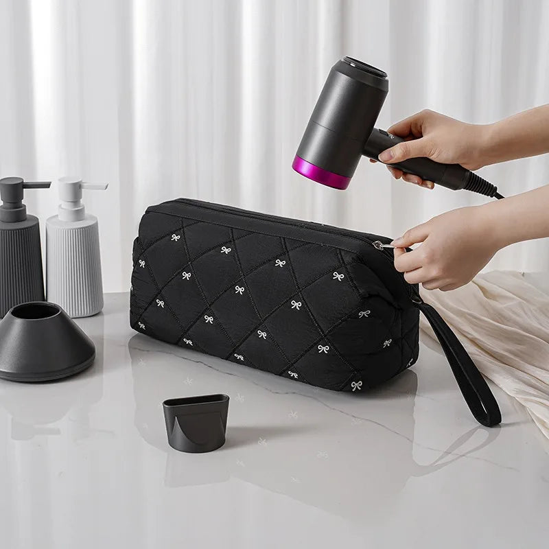 A hand is placing a portable hair dryer and styling tool into a black quilted storage bag on a white surface.