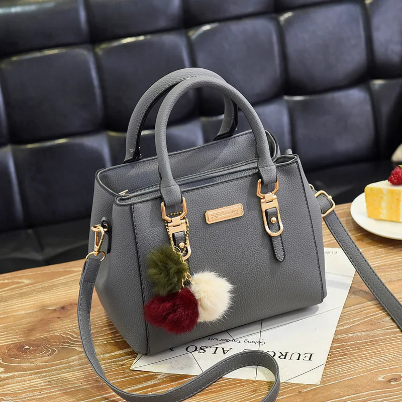 A gray vintage PU leather shoulder bag with gold hardware and a fluffy pom-pom charm resting on a wooden surface.