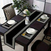 A set of black geometric table runners and placemats with gold accents on a dark wooden table.