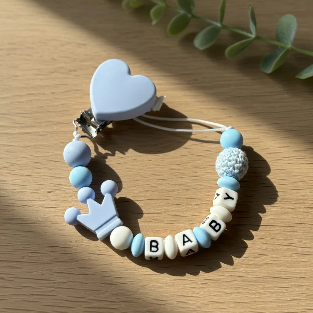 A light blue pacifier clip bracelet featuring a heart charm and custom name beads on a wooden surface.