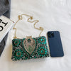 A green and gold rhinestone evening clutch with a gold chain strap resting on a white surface next to a smartphone.