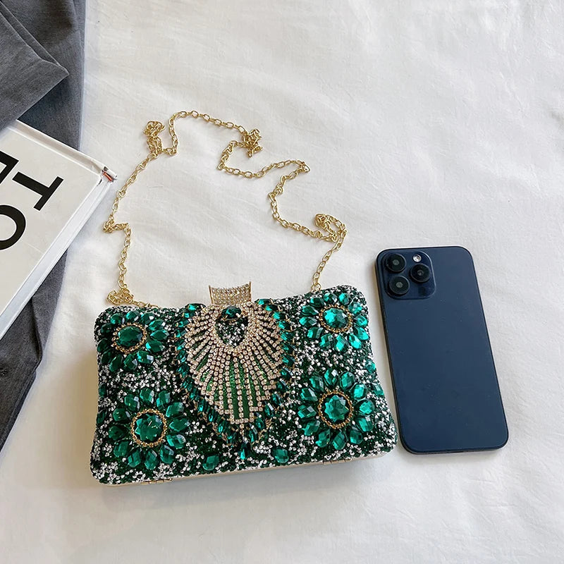 A green and gold rhinestone evening clutch with a gold chain strap resting on a white surface next to a smartphone.